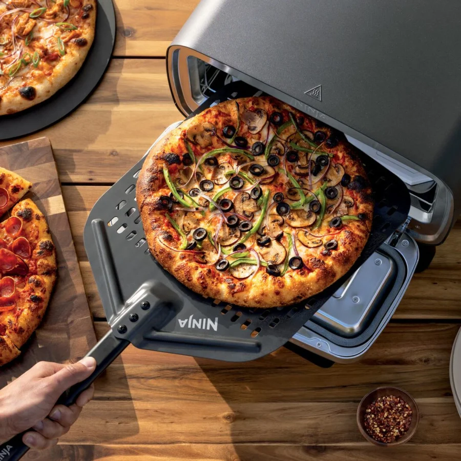 Une pizza sortie du Ninja Artisan avec sa pelle perforée, d’autres pizzas sur la table