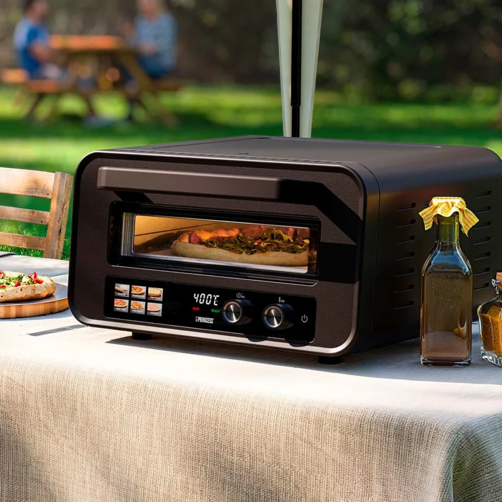 Princess 112780 sur une table de jardin, une pizza en cuisson derrière la vitre, une autre servie à côté
