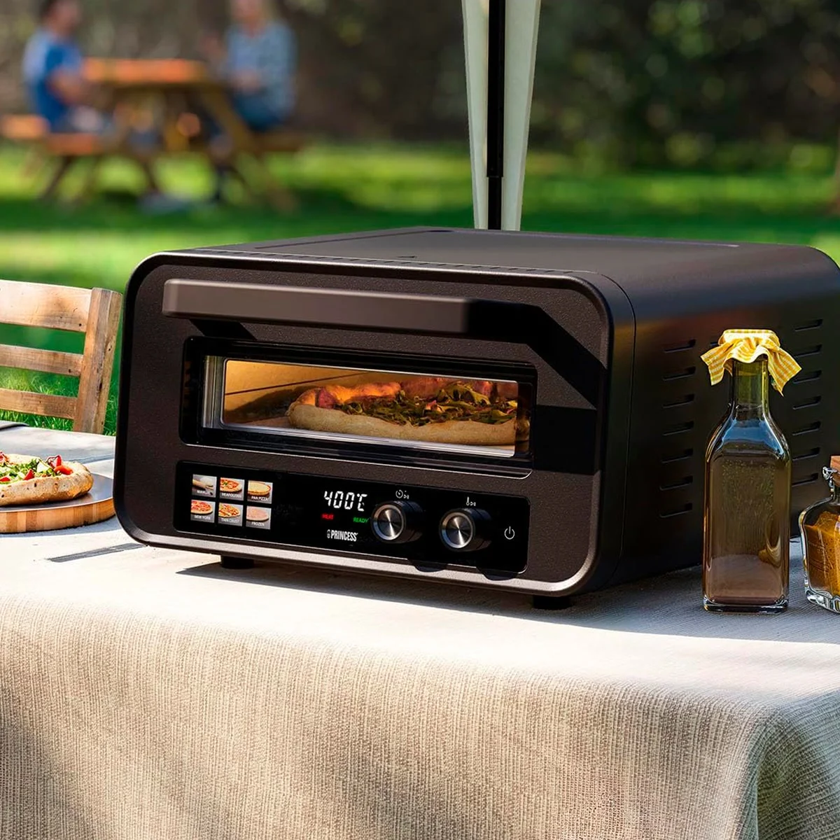 Princess 112780 sur une table de jardin, une pizza en cuisson derrière la vitre, une autre servie à côté