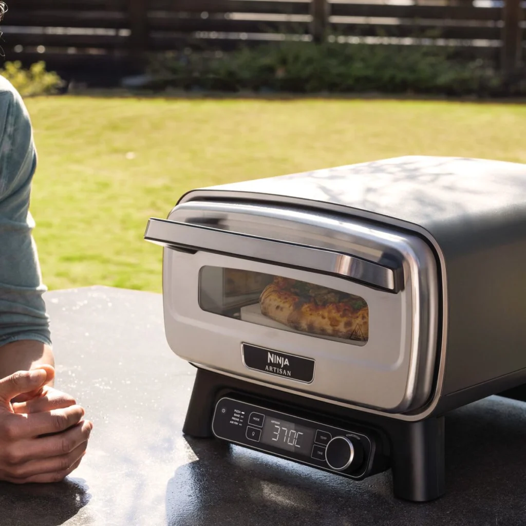 Le Ninja Artisan dans un jardin : la pizza cuit derrière la fenêtre éclairée