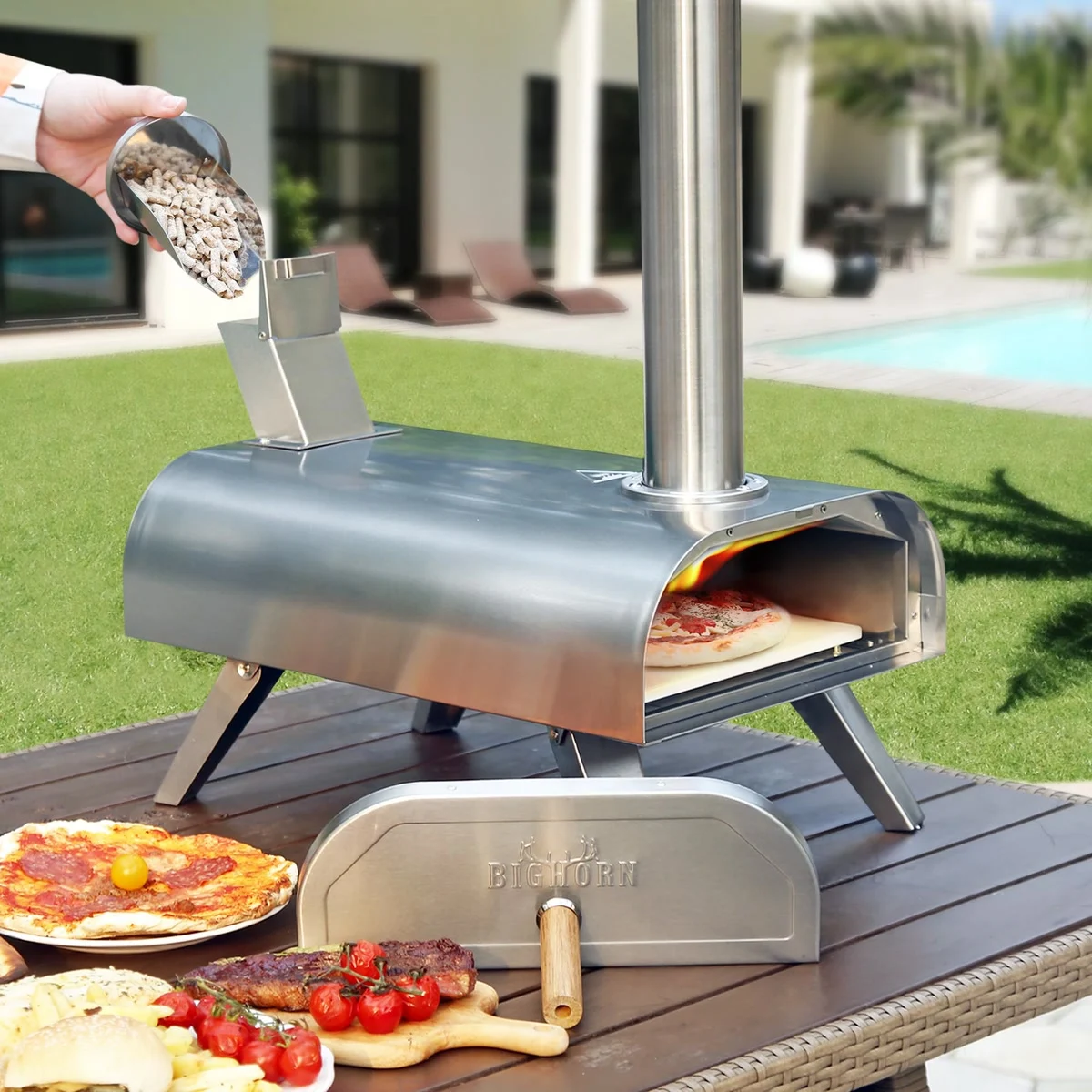 Une main verse des granulés de bois dans la trémie du Bighorn 12", une pizza en cuisson, au bord d’une piscine