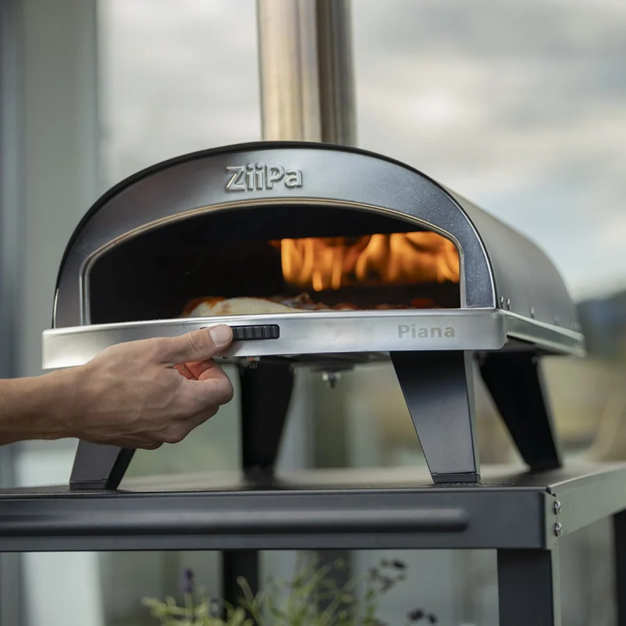 Une main tourne la molette de la pierre rotative du ZiiPa Piana, une pizza en cuisson devant les flammes