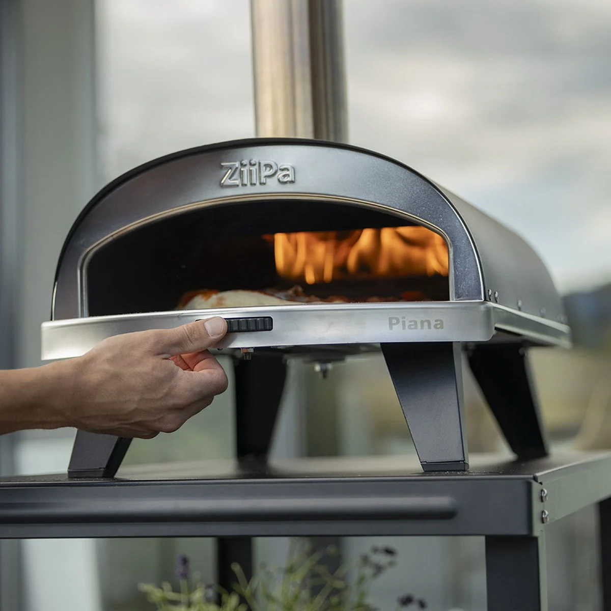 Une main tourne la molette de la pierre rotative du ZiiPa Piana, une pizza en cuisson devant les flammes