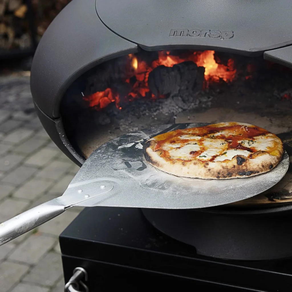 Une pizza sort du Morsø Forno sur une pelle (non fournie), les braises au fond