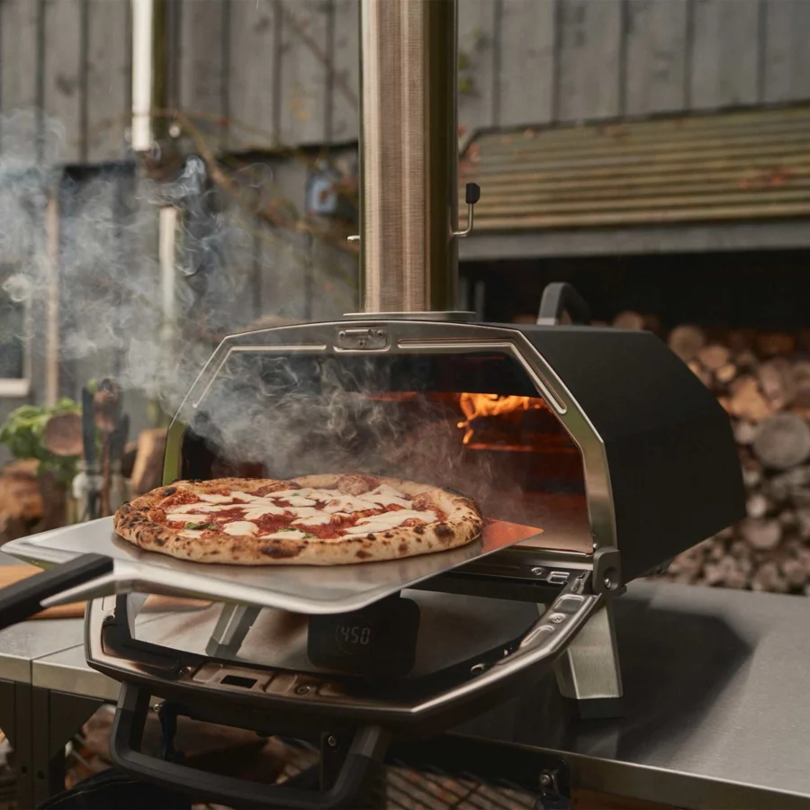 Une pizza margherita sort de l’Ooni Karu 2 Pro sur une pelle, porte ouverte, bois en arrière-plan