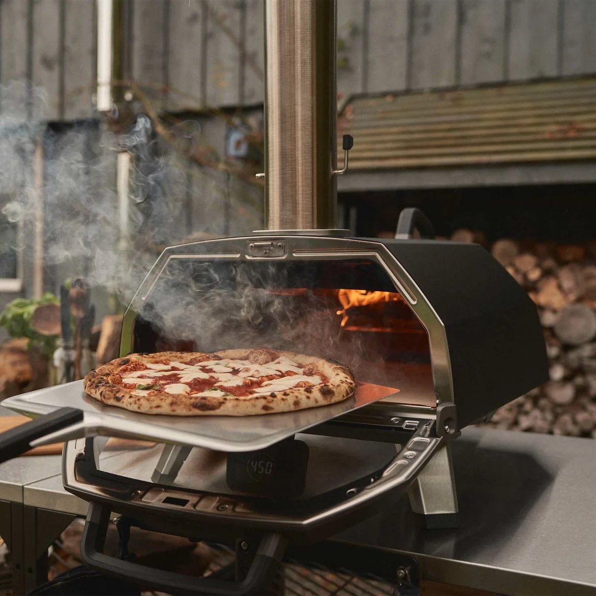 Une pizza margherita sort de l’Ooni Karu 2 Pro sur une pelle, porte ouverte, bois en arrière-plan