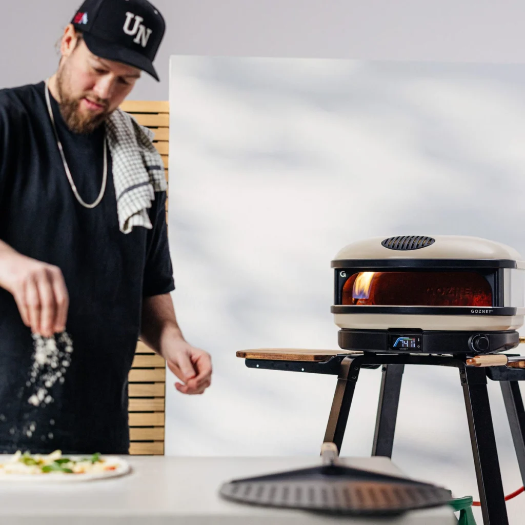 Un pizzaïolo garnit une pizza, le Gozney Arc XL crème allumé sur son support