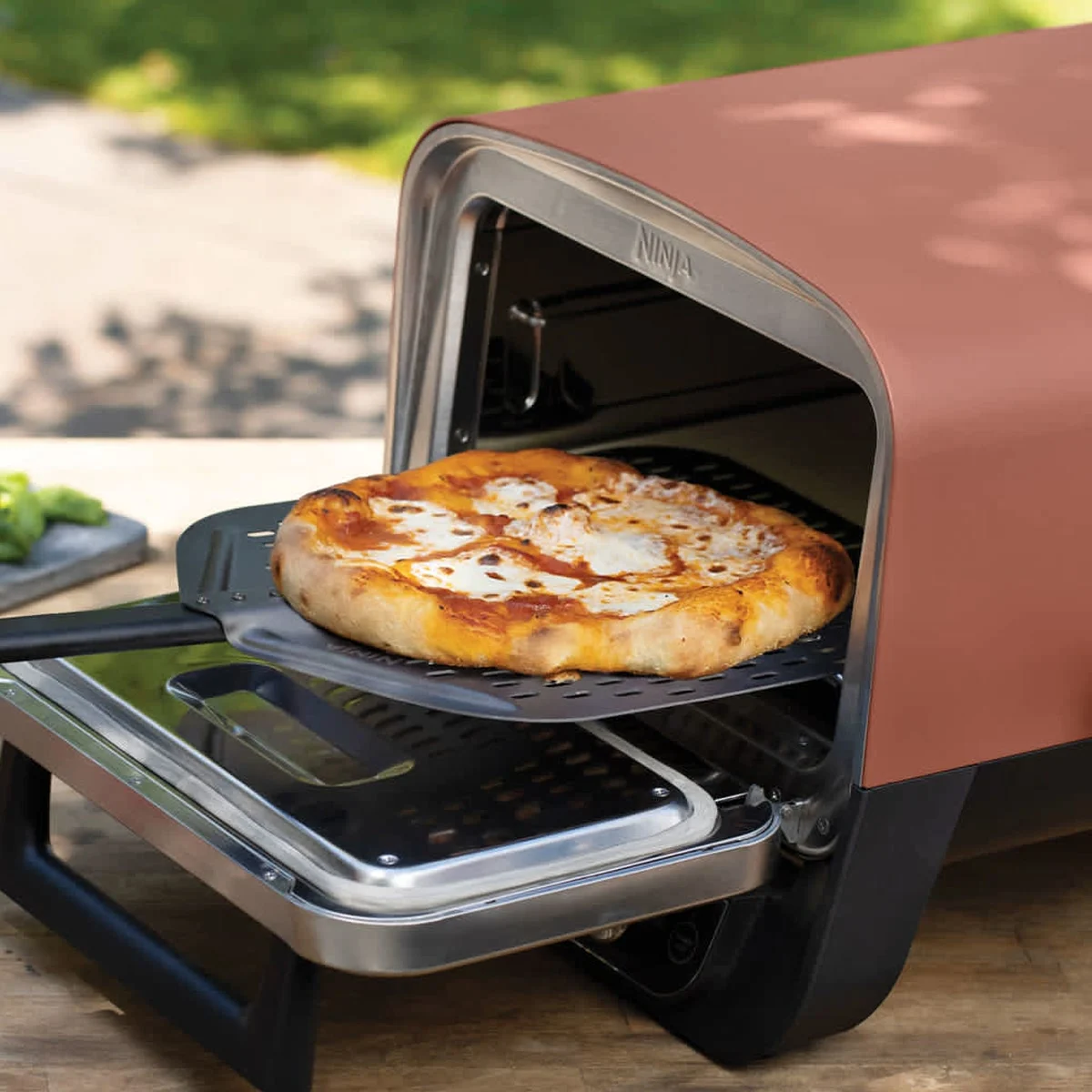 Une pizza sortie à la pelle du Ninja Woodfire, porte ouverte, sur une table de jardin