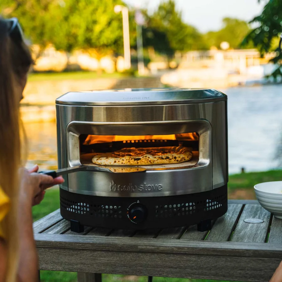 Deux amies font cuire une pizza dans le Solo Stove Pi Prime posé sur une table en bois, au bord de l’eau