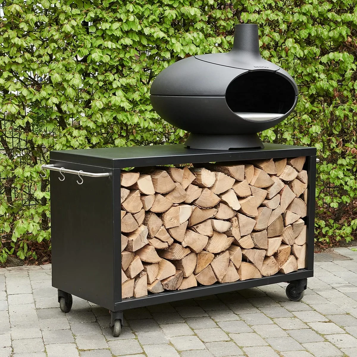 Morsø Forno sur une table roulante remplie de bûches (vendue à part), devant une haie